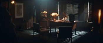 Movie still from “Archenemy” (2020), directed by Adam Egypt Mortimer – A man sitting at a desk in front of two lamps; Wide shot, Over the shoulder angle