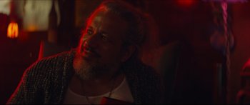 Movie still from “Archenemy” (2020), directed by Adam Egypt Mortimer – A man with long hair and a beard is smiling for the camera; Close Up shot, Over the shoulder angle