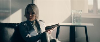 Movie still from “Archenemy” (2020), directed by Adam Egypt Mortimer – A woman holding a gun while sitting on a chair; Medium shot, Low angle