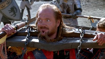 Movie still from “Army of Darkness” (1992), directed by Sam Raimi – A man with long hair and a beard is sitting in chains; Close Up shot, Over the shoulder angle