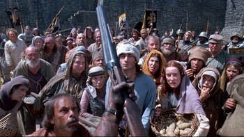 Movie still from “Army of Darkness” (1992), directed by Sam Raimi – A group of people standing around a man holding a rifle; Wide shot, High angle