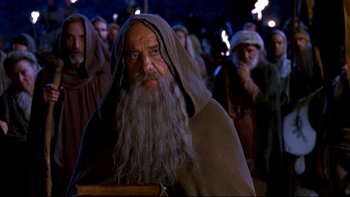 Movie still from “Army of Darkness” (1992), directed by Sam Raimi – An old man with a long beard is in a crowd of people; Medium shot, Over the shoulder angle