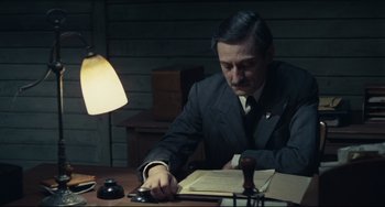 Movie still from “Army of Shadows” (1969), directed by Jean-Pierre Melville – A man in a suit and tie sitting at a desk writing; Medium shot, High angle