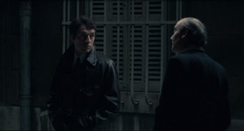 Movie still from “Army of Shadows” (1969), directed by Jean-Pierre Melville – Two men standing next to each other in front of a metal gate; Medium shot, Over the shoulder angle