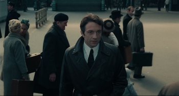 Movie still from “Army of Shadows” (1969), directed by Jean-Pierre Melville – A man in a black coat and tie standing next to a crowd; Medium shot, Low angle