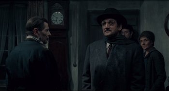 Movie still from “Army of Shadows” (1969), directed by Jean-Pierre Melville – A man wearing a hat and a suit and tie standing in front of a grandfather clock; Medium shot, Over the shoulder angle