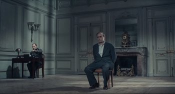 Movie still from “Army of Shadows” (1969), directed by Jean-Pierre Melville – A man in a suit sitting on a chair in front of a fireplace; Wide shot, High angle