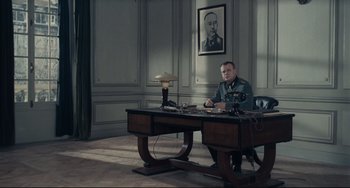 Movie still from “Army of Shadows” (1969), directed by Jean-Pierre Melville – A man sitting at a desk in front of a picture; Wide shot, Low angle