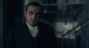 Movie still from “Army of Shadows” (1969), directed by Jean-Pierre Melville – A man in a suit and glasses is standing in a room; Medium shot, Over the shoulder angle