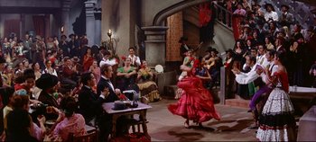 Movie still from “Around the World in 80 Days” (1956), directed by John Farrow – A group of people sitting around a room with a woman in a red dress dancing; Wide shot, High angle