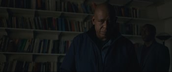 Movie still from “Arrival” (2016), directed by Denis Villeneuve – A bald man in a jacket is in front of a book shelf; Medium shot, Low angle