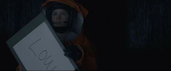 Movie still from “Arrival” (2016), directed by Denis Villeneuve – A person in a space suit holding an object in front of a dark background; Close Up shot, Overhead angle