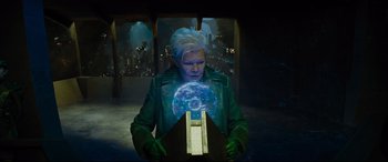 Movie still from “Artemis Fowl” (2020), directed by Kenneth Branagh – A man in a green jacket is holding an object in front of a crowd; Medium shot, Over the shoulder angle