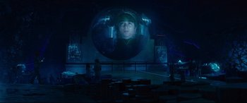 Movie still from “Artemis Fowl” (2020), directed by Kenneth Branagh – A large projection of a man's face on a wall in a dark room; Wide shot, Over the shoulder angle