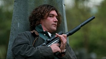 Movie still from “Assassins” (1995), directed by Richard Donner – A man holding a gun while standing next to a tree; Close Up shot, Low angle