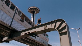 Movie still from “Assassins” (1995), directed by Richard Donner – A man running across a bridge near a train; Extreme Wide shot, Low angle