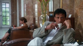 Movie still from “Assassins” (1995), directed by Richard Donner – A man sitting in a chair drinking a cup of coffee; Medium shot, Low angle