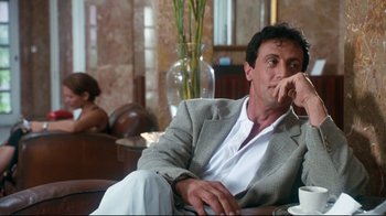 Movie still from “Assassins” (1995), directed by Richard Donner – A man sitting on a couch in a living room; Medium shot, Over the shoulder angle