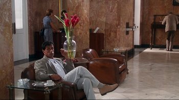 Movie still from “Assassins” (1995), directed by Richard Donner – A man sitting on a chair in a room; Wide shot, Low angle