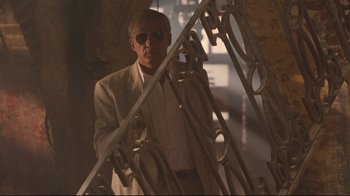 Movie still from “Assassins” (1995), directed by Richard Donner – A man in a suit and sunglasses standing next to a metal structure; Medium shot, Low angle