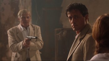 Movie still from “Assassins” (1995), directed by Richard Donner – A man in a suit and tie holding a gun in front of another man in a suit and tie; Medium shot, Over the shoulder angle