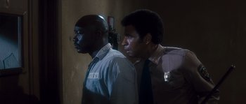 Movie still from “Assault on Precinct 13” (1976), directed by John Carpenter – Two police officers standing next to each other in a dark room; Medium shot, Over the shoulder angle