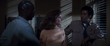 Movie still from “Assault on Precinct 13” (1976), directed by John Carpenter – A woman with a scar on her arm is being held by a police officer; Medium shot, Over the shoulder angle
