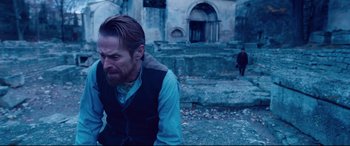 Movie still from “At Eternity's Gate” (2018), directed by Julian Schnabel – A man sitting on the ground in front of a stone building; Medium shot, Over the shoulder angle
