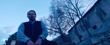 Movie still from “At Eternity's Gate” (2018), directed by Julian Schnabel – A man sitting on the side of a building next to a tree; Medium shot, Low angle