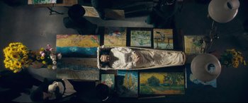 Movie still from “At Eternity's Gate” (2018), directed by Julian Schnabel – A person in a casket surrounded by paintings; Wide shot, Overhead angle
