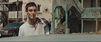 Movie still from “At First Light” (2018), directed by Jason Stone – A young man smoking a cigarette while sitting on a bench; Medium shot, Over the shoulder angle