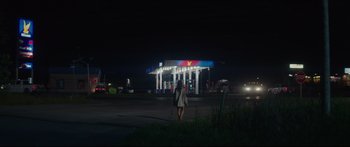 Movie still from “At First Light” (2018), directed by Jason Stone – A woman is standing in a parking lot near a gas station at night; Extreme Wide shot, Low angle