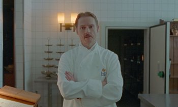Movie still from “Spencer” (2021), directed by Pablo Larraín – A man standing in a kitchen with his arms crossed; Medium shot, Low angle