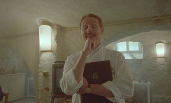 Movie still from “Spencer” (2021), directed by Pablo Larraín – A man holding a book in his hand and posing for a picture; Medium shot, Low angle