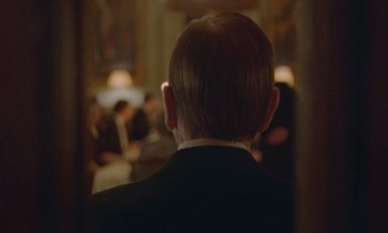 Movie still from “Spencer” (2021), directed by Pablo Larraín – A man in a black suit is looking at a group of people; Close Up shot, Over the shoulder angle