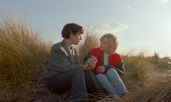 Movie still from “Spencer” (2021), directed by Pablo Larraín – A man and a woman sitting in the grass holding hands; Medium shot, High angle