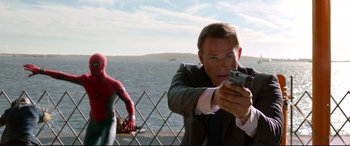 Movie still from “Spider-Man: Homecoming” (2017), directed by Jon Watts – A man in a suit holding a gun in front of water; Medium shot, Over the shoulder angle