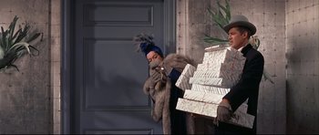 Movie still from “Auntie Mame” (1958), directed by Morton DaCosta – A man in a blue hat and a fur coat holding a stack of boxes; Medium shot, High angle
