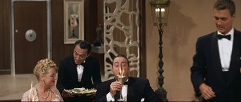 Movie still from “Auntie Mame” (1958), directed by Morton DaCosta – A man in a suit and tie lighting a candle; Medium shot, Over the shoulder angle