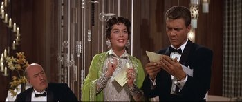 Movie still from “Auntie Mame” (1958), directed by Morton DaCosta – A woman holding a piece of paper while standing next to a man; Medium shot, Over the shoulder angle