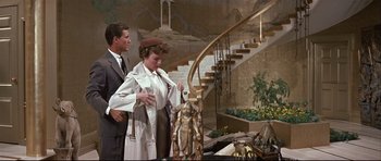 Movie still from “Auntie Mame” (1958), directed by Morton DaCosta – A man and a woman standing next to each other in front of a staircase; Wide shot, High angle