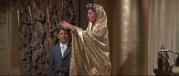 Movie still from “Auntie Mame” (1958), directed by Morton DaCosta – An older woman in a gold dress is holding a child in front of him; Medium shot, Over the shoulder angle