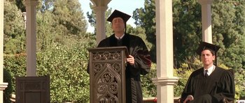 Movie still from “Austin Powers in Goldmember” (2002), directed by Jay Roach – A man wearing a graduation cap and gown standing at a podium; Medium shot, Low angle