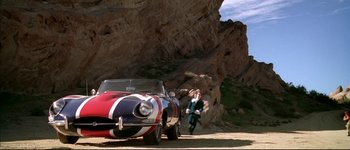 Movie still from “Austin Powers: International Man of Mystery” (1997), directed by Jay Roach – A man standing next to a car on the side of a mountain; Extreme Wide shot, High angle