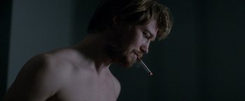 Movie still from “Stars at Noon” (2022), directed by Claire Denis – A shirtless man smoking a cigarette in a dark room; Close Up shot, Over the shoulder angle