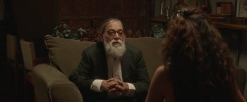 Movie still from “Stars at Noon” (2022), directed by Claire Denis – An older man sitting on a couch talking to a woman; Medium shot, Over the shoulder angle