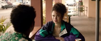 Movie still from “Stars at Noon” (2022), directed by Claire Denis – A young woman sitting on a bench looking at the camera; Close Up shot, Over the shoulder angle