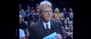 Movie still from “Austin Powers: The Spy Who Shagged Me” (1999), directed by Jay Roach – A man in a suit and tie holding a microphone in front of an audience; Medium shot, Over the shoulder angle