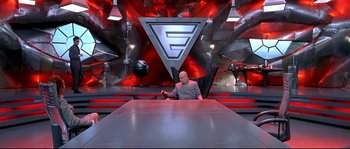 Movie still from “Austin Powers: The Spy Who Shagged Me” (1999), directed by Jay Roach – A man sitting at a table in front of a red background; Wide shot, High angle