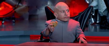 Movie still from “Austin Powers: The Spy Who Shagged Me” (1999), directed by Jay Roach – A bald man sitting at a table holding a piece of food; Medium shot, Low angle